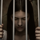 Lianne casts down her eyes while holding onto the bars of the cradle she's trapped inn.
